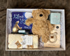 Gift box with teddy bear, blanket, and baby items on a textured surface