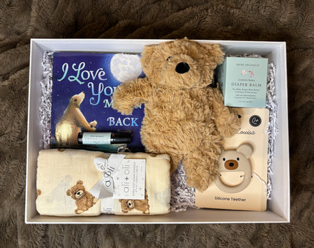 Gift box with teddy bear, blanket, and baby items on a textured surface