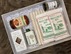 Gourmet pizza date night gift box featuring Rao’s Margherita pizza sauce, Weisenberger pizza crust mixes, garlic-infused olive oil, Rustic Bakery gingerbread cookies, an Heraclea olive oil tin, a jar of herbs de Provence seasoning, and a striped kitchen towel arranged in a white gift box with crinkle paper.