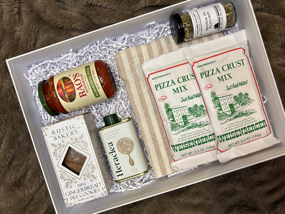Gourmet pizza date night gift box featuring Rao’s Margherita pizza sauce, Weisenberger pizza crust mixes, garlic-infused olive oil, Rustic Bakery gingerbread cookies, an Heraclea olive oil tin, a jar of herbs de Provence seasoning, and a striped kitchen towel arranged in a white gift box with crinkle paper.