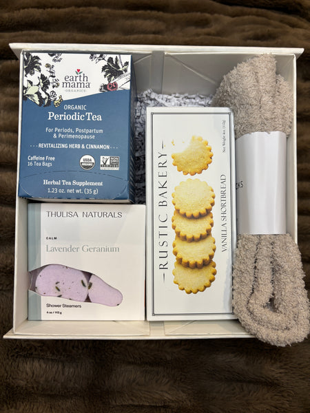 Curated period care gift set including herbal tea, sweet shortbread cookies, soothing shower steamers, plush socks, and a body scrub styled in a box on a textured surface for period comfort and relaxation.