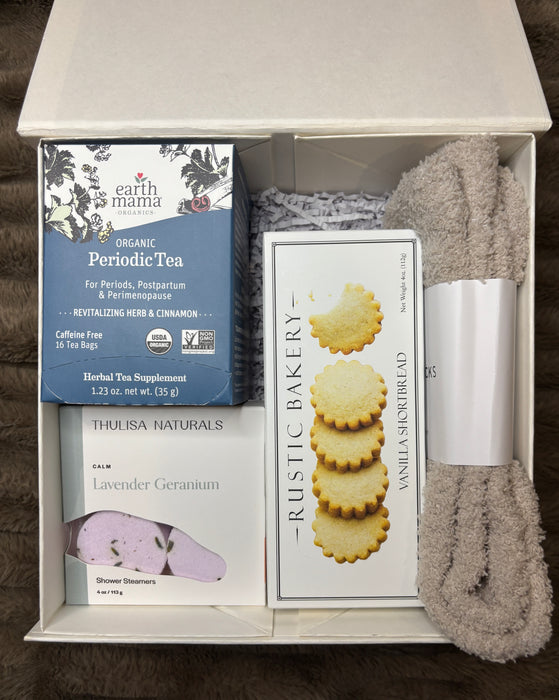 Comfort-focused period support box with tea, cookies, calming shower steamers, cloud socks, and a bath scrub displayed together on a textured background.