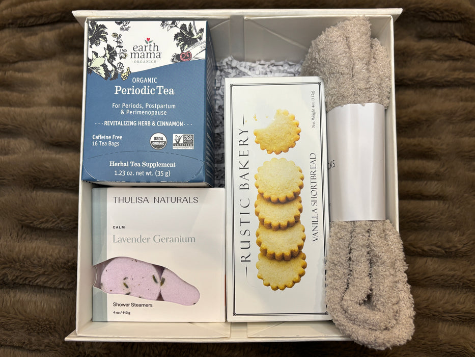 Period support gift box featuring Organic Periodic Tea, vanilla bean shortbread cookies, lavender-geranium shower steamers, a body scrub, and cozy socks arranged neatly on a textured surface.