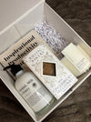 Gift set with skincare products and a book inside a box on a textured surface. Rest & Recharge.